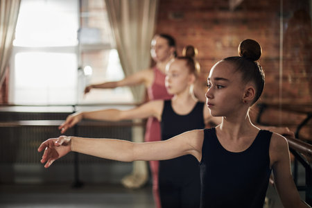 Teenage girls practicing ballet positions at barre in dance studio with female instructor guiding in...