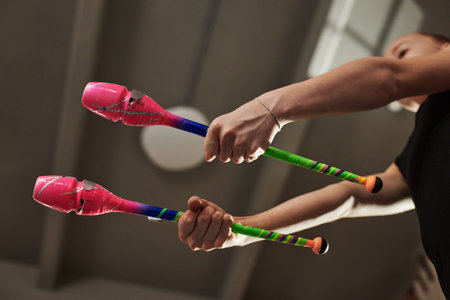 Teenage girl holding two colorful rhythmic gymnastics clubs, performing routine indoors with arms...