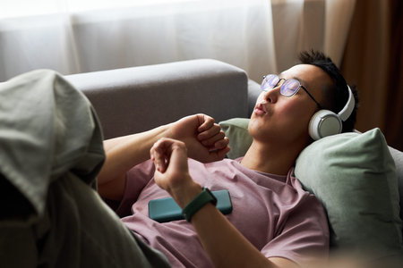 Young adult man relaxing on sofa listening to music with wireless headphones, eyes closed, holding...