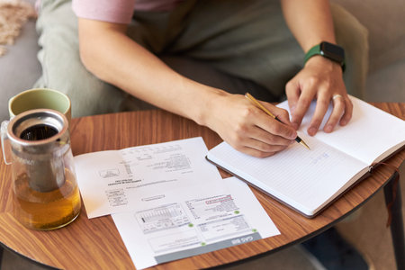 Young adult Asian man sitting at table writing in notebook with pen while reviewing utility bills...
