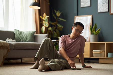 Asian young adult man sitting on floor in living room using smartphone, leaning back with relaxed posture, natural light coming through window, casual home environmentの写真素材