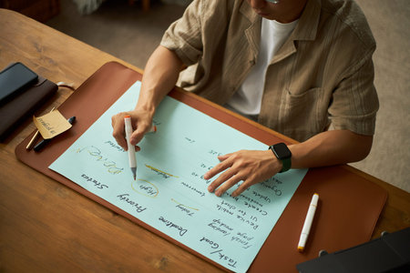 Young adult Asian man sitting at desk writing on large sheet of paper with marker, planning project tasks and priorities, using smartwatch and digital tablet for organizationの写真素材