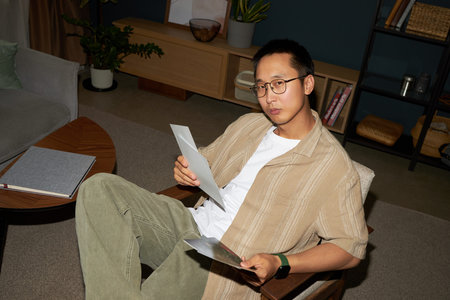 Portrait of young adult Asian man sitting in modern living room holding documents and looking into...