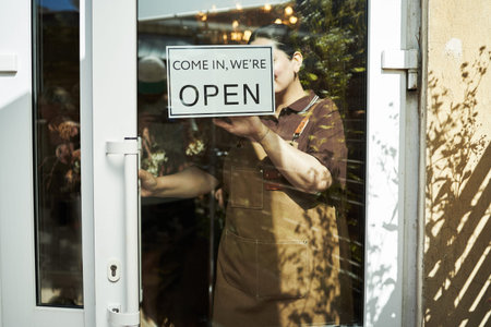 Caucasian middle aged woman opening glass door of shop while placing open sign on entrance, standing inside storefront, partially visible face, wearing apron, preparing for businessの写真素材