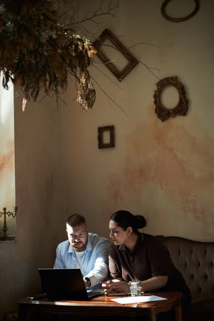 Young adult man and young adult woman collaborating at table using laptop, both focused on screen,...
