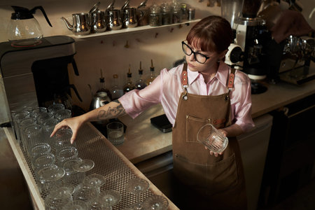 Caucasian young adult woman with glasses arranging clean glassware on counter in coffee shop,...