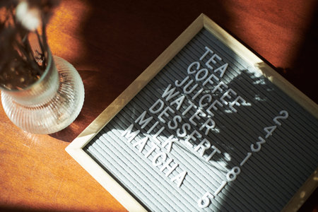 Letter board displaying words tea, coffee, juice, water, dessert, milk, matcha and numbers on wooden table, sunlight casting shadows across surface, glass vase partially visibleの写真素材