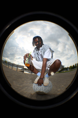 Fisheye lens view of young man crouching on stone outdoors holding plastic cup with straw, looking...