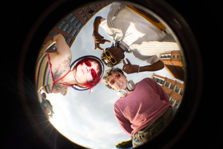 Group of three teenagers, including Caucasian girl with tattoos, Black boy making hand gesture, and hipster boy with headphones, standing outdoors looking down at cameraの写真素材