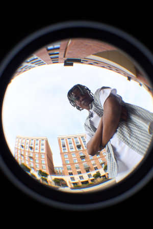Black teenager boy standing outdoors with arms crossed, looking down at camera through fisheye lens,...