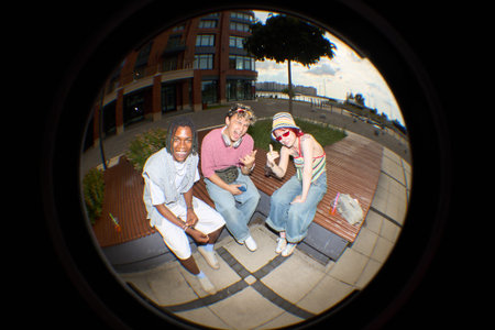 Group of three teenagers, Black, Caucasian male, female, sitting on outdoor bench, smiling and...