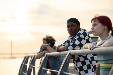 Group of multiethnic teenagers leaning on metal railing by waterfront, Black teenage boy looking at...