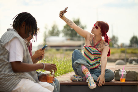 Young woman with tattoos sitting outdoors taking selfie with smartphone while man next to her using...