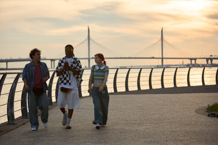 Group of three teenagers, two Caucasian males and one female, walking along waterfront promenade at...