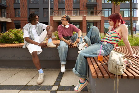 Group of three teenagers, Black, Caucasian male, female, sitting on outdoor bench, talking and relaxing together, headphones and drink cup visible, urban backgroundの写真素材