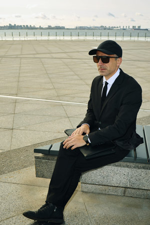 Caucasian businessman wearing suit, sunglasses and black cap sitting on outdoor bench holding...