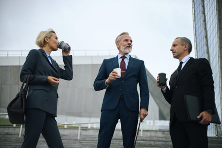 Three middle aged Caucasian business professionals standing outdoors, holding disposable coffee...