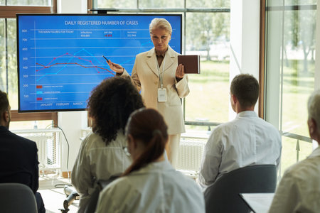 Caucasian female medical clinic executive presenting data on large screen to diverse group of young...