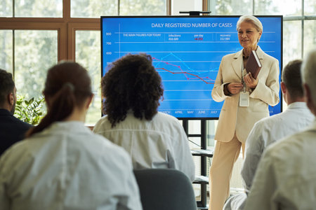 Senior Caucasian woman presenting healthcare medical statistics on large screen to group of diverse...