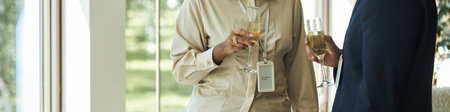 Caucasian middle aged woman and Black middle aged man holding champagne glasses and talking near...