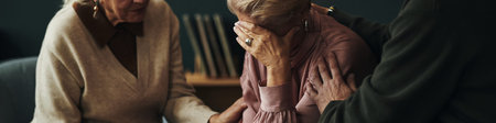 Header of senior woman sitting with head in hand, appearing distressed, while being comforted by female and male couple of friends, placing hands on her shouldersの写真素材