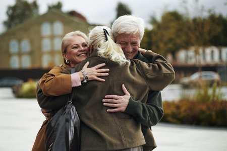 Group of three senior Caucasian people embracing outdoors, two women and one man smiling warmly while hugging each other, standing in urban park setting with blurred buildingsの写真素材