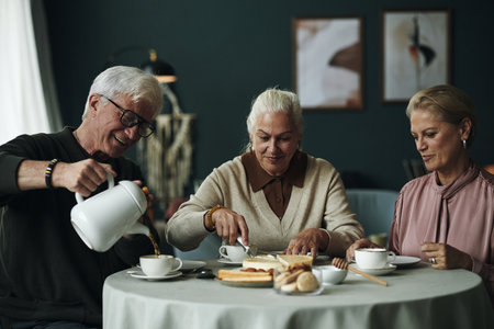 Three Caucasian seniors sitting at round table enjoying dessert and coffee together, senior man pouring coffee while two female friends cutting and serving cake, smiling and interactingの写真素材