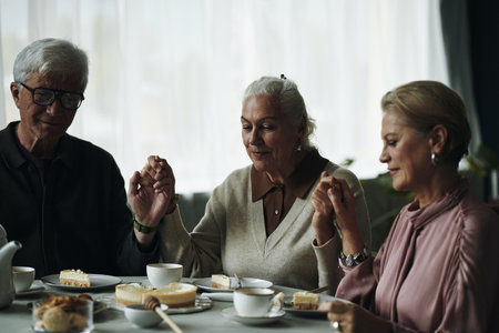 Senior Caucasian man and two elderly women friends sitting at table holding hands before eating dessert, engaging in moment of gratitude or prayer, sharing time together indoorsの写真素材