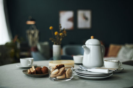Table set with glass bowl of assorted cookies, plates with napkins, cups, and ceramic teapot arranged for serving snacks and hot beverages in cozy indoor settingの写真素材