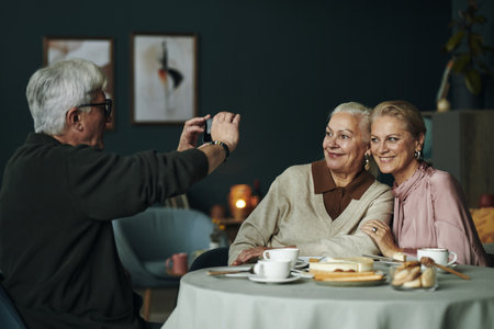 Senior Caucasian man photographing senior woman and middle aged female friend sitting together at table smiling, enjoying dessert and drinks in cozy indoor settingの写真素材