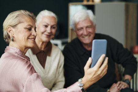 Senior Caucasian woman taking selfie with smartphone while sitting with male and female friend...