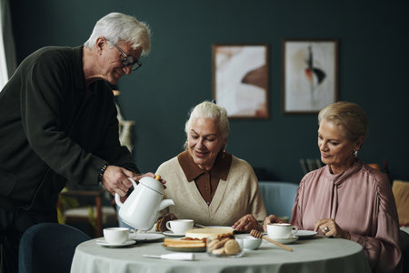 Senior Caucasian man pouring tea for two elderly women sitting at round table enjoying conversation and desserts in cozy indoor setting, all friends smiling and engagedの写真素材