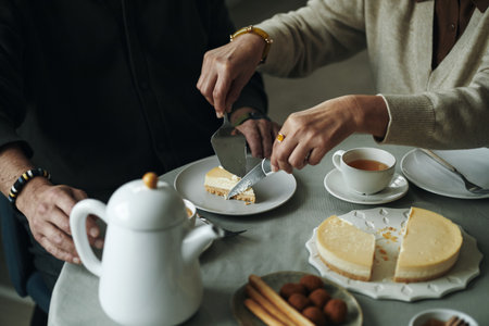 Senior Caucasian woman cutting cheesecake with cake server while male friend or husband sitting beside her at table, hands visible, sharing dessert during tea time, teapot and cup presentの写真素材