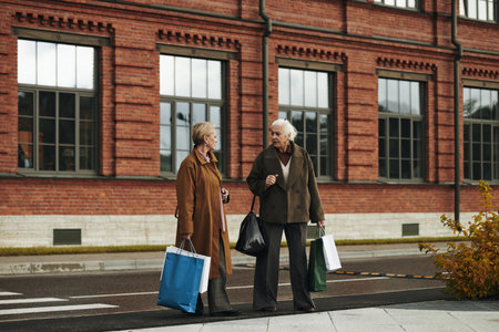 Two senior Caucasian women walking outdoors carrying shopping bags, engaging in conversation on city street with brick building in background, both appearing relaxed and focusedの写真素材