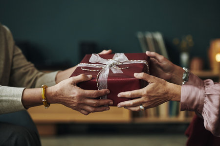 Hands of senior woman handing wrapped gift box to elderly female friend, girlfriends exchanging present, focus on gesture of giving and receiving, home setting visibleの写真素材