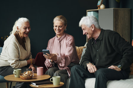Three senior Caucasian people sitting together, smiling and looking at smartphone, two women and one man engaging in conversation, sharing moment in casual indoor settingの写真素材