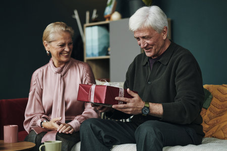 Senior Caucasian man sitting on sofa opening gift box while elderly wife is smiling and watching him, both appearing joyful and engaged in living room settingの写真素材