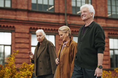 Group of senior Caucasian man and two women in warm coats walking outdoors in front of brick building, smiling and talking, enjoying time together during autumn dayの写真素材