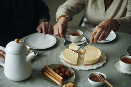 Hands of senior woman slicing cheesecake while having tea with friends sitting beside her at table...