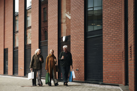 Two senior Caucasian women and man walking together outdoors carrying shopping bags and coffee cups, friends smiling and talking while strolling along modern brick building in urban settingの写真素材