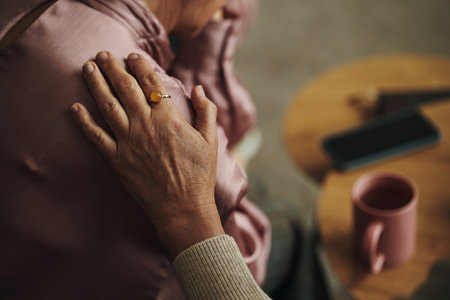 Elderly woman comforting senior female friend by gently placing hand on shoulder, woman sitting with head resting on hand near smartphone and coffee mug in living roomの写真素材