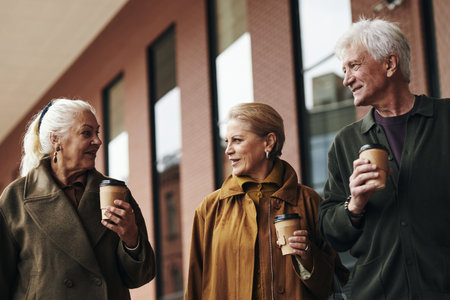 Two elderly Caucasian women and senior man walking outdoors holding coffee cups, engaging in conversation, smiling at each other, standing in front of modern building, casual interactionの写真素材