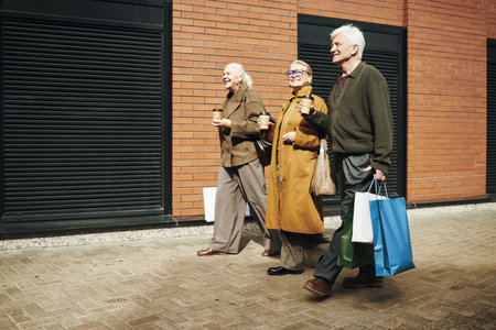 Three senior Caucasian people walking outdoors carrying shopping bags and takeaway coffee cups, smiling and talking together in urban setting near brick buildingの写真素材