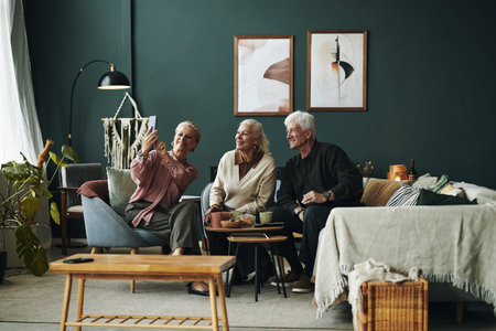 Three senior male and female friends sitting together on sofa smiling and taking selfie with smartphone in modern living room, enjoying leisure time and socializing indoorsの写真素材