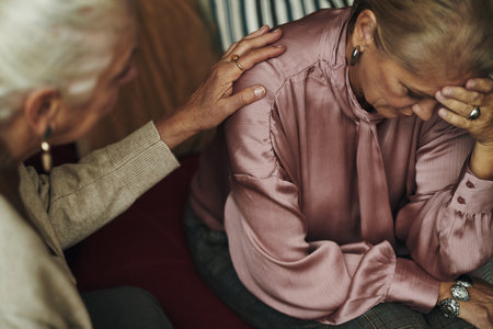 Senior Caucasian woman sitting with head in hand appearing distressed while female friend comforting her by gently placing hand on her shoulder indoorsの写真素材