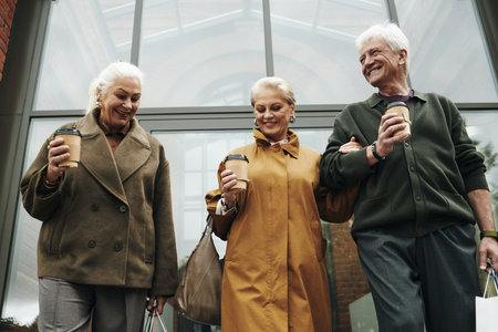 Three senior Caucasian people walking outdoors holding takeaway coffee cups and shopping bags, two women and one man smiling and interacting while strolling togetherの写真素材