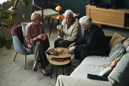 Three senior Caucasian women and man sitting together in living room drinking hot beverages and...