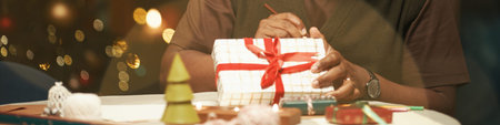 Middle aged Black man wrapping Christmas gift box with red ribbon sitting at table, hands tying bow, festive decorations and Christmas tree lights visible in backgroundの写真素材