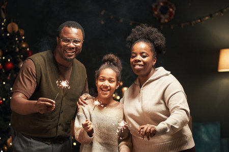 Black man, Black woman, and Black child smiling and holding sparklers together in festive room with...