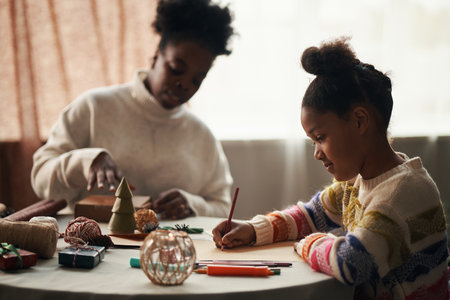 Black girl drawing with colored pencils at table while Black woman wrapping gifts in background, both engaging in Christmas holiday activities with festive decorations and craft suppliesの写真素材